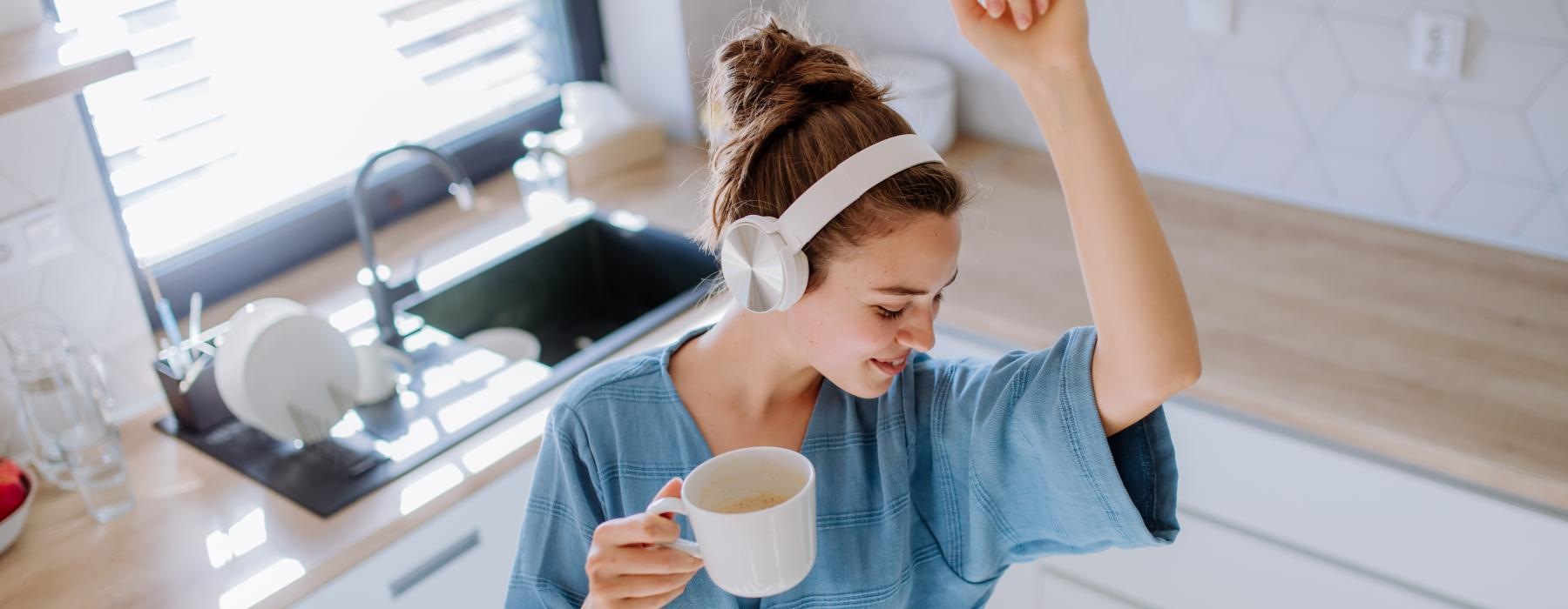 woman with headphones dances in her kitchen while holding a cup of coffee